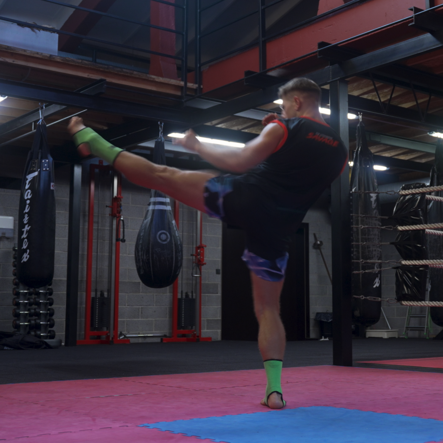 Person practicing boxing in a gym with punching bags and boxing ring in the background.