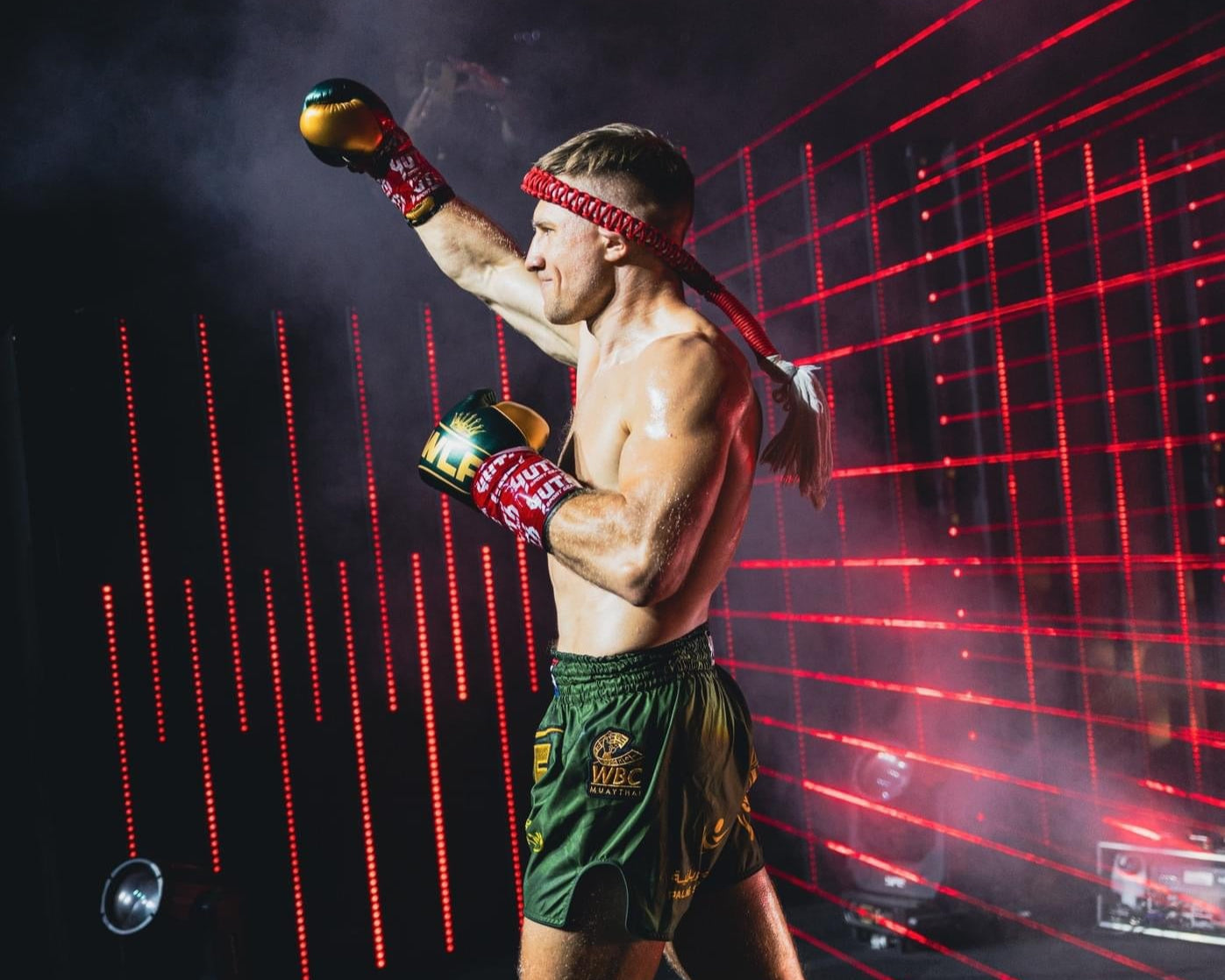 Boxer in green shorts celebrating with raised fists against a dark background with red lights.