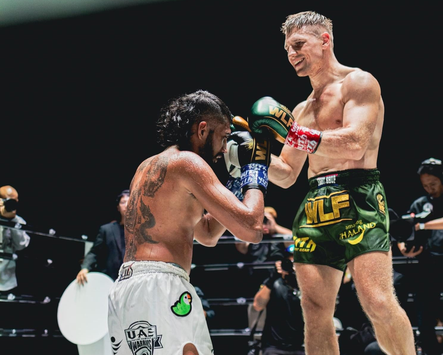 Two fighters on a boxing ring with one on his knees and the other standing, surrounded by photographers.